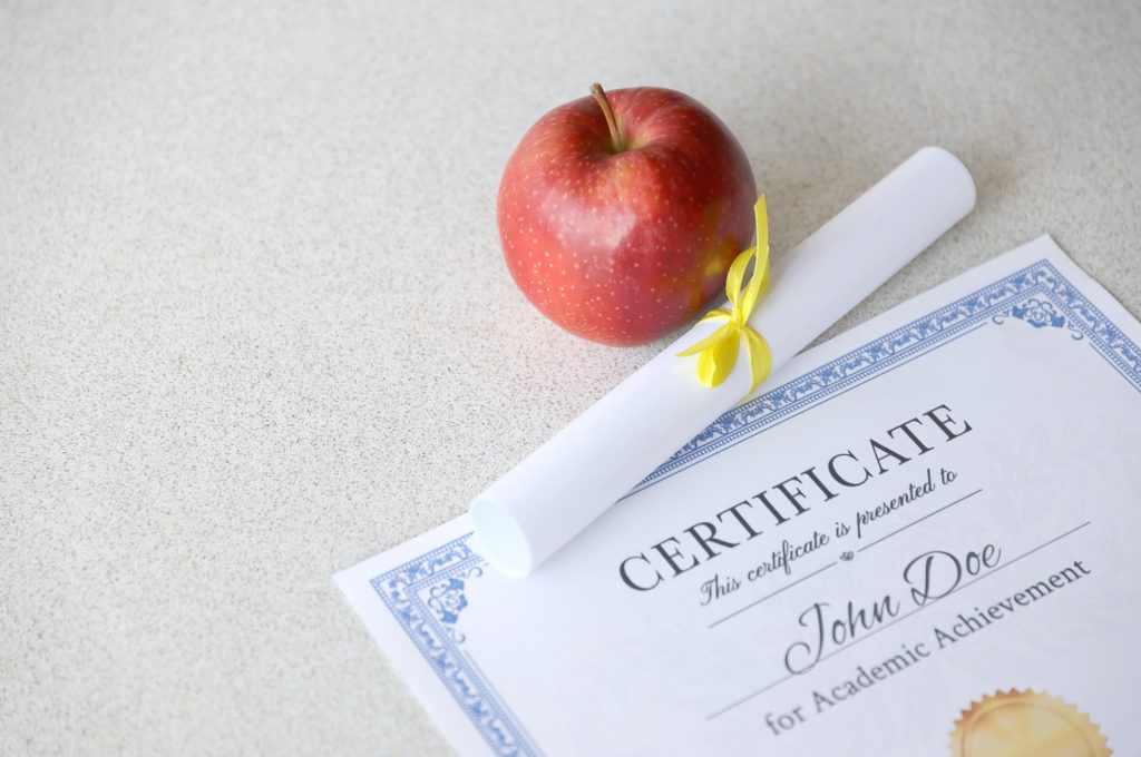 A certificate of achievement lies on table with small scroll and red apple. Education documents